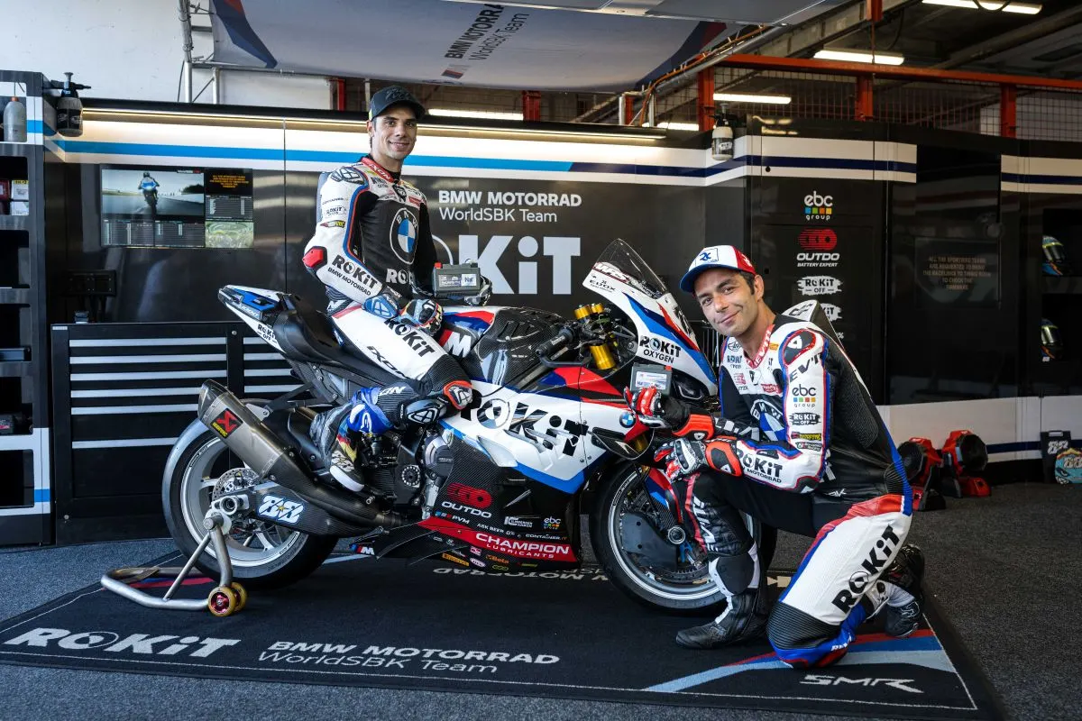 One BMW Motorrad WorldSBK rider seated on the race bike and one kneeling beside it, each holding an OUTDO sodium-ion battery